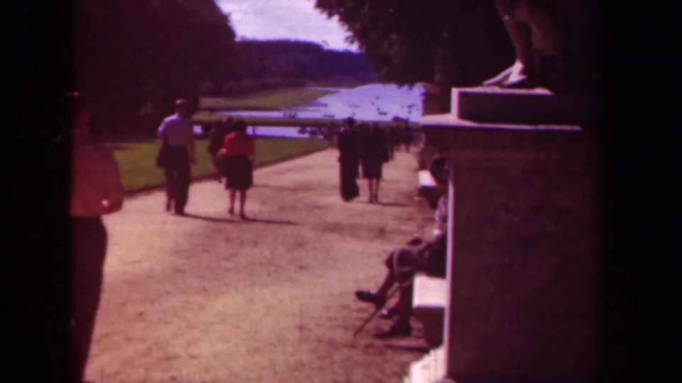 A handheld 8mm view shows a visitor entering the frame as others stroll through a formal Paris park landscape.