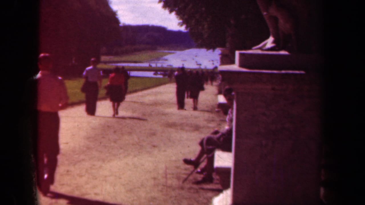 People walk along a broad park path beside lawns, trees, a pond, and a bench.