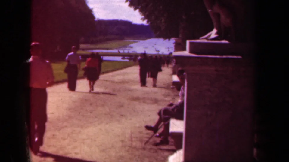 1939 Paris park view with adults walking near a pond, a seated visitor on a bench, lawns, and tall trees.