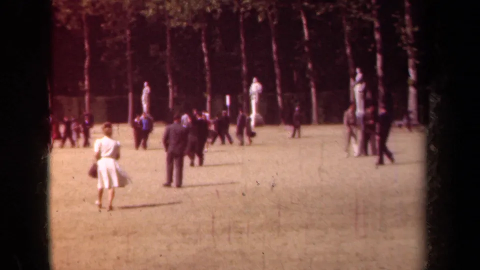 1939 Paris park lawn with visitors in suits and dresses walking among classical statues and trees.