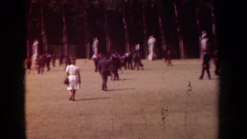 1939 Paris park scene with a woman in a white dress walking toward groups near statues and trees.