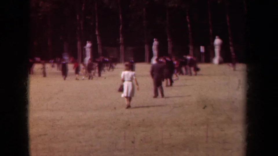 1939 Paris park lawn with adults strolling and gathering near classical statues under tall trees.