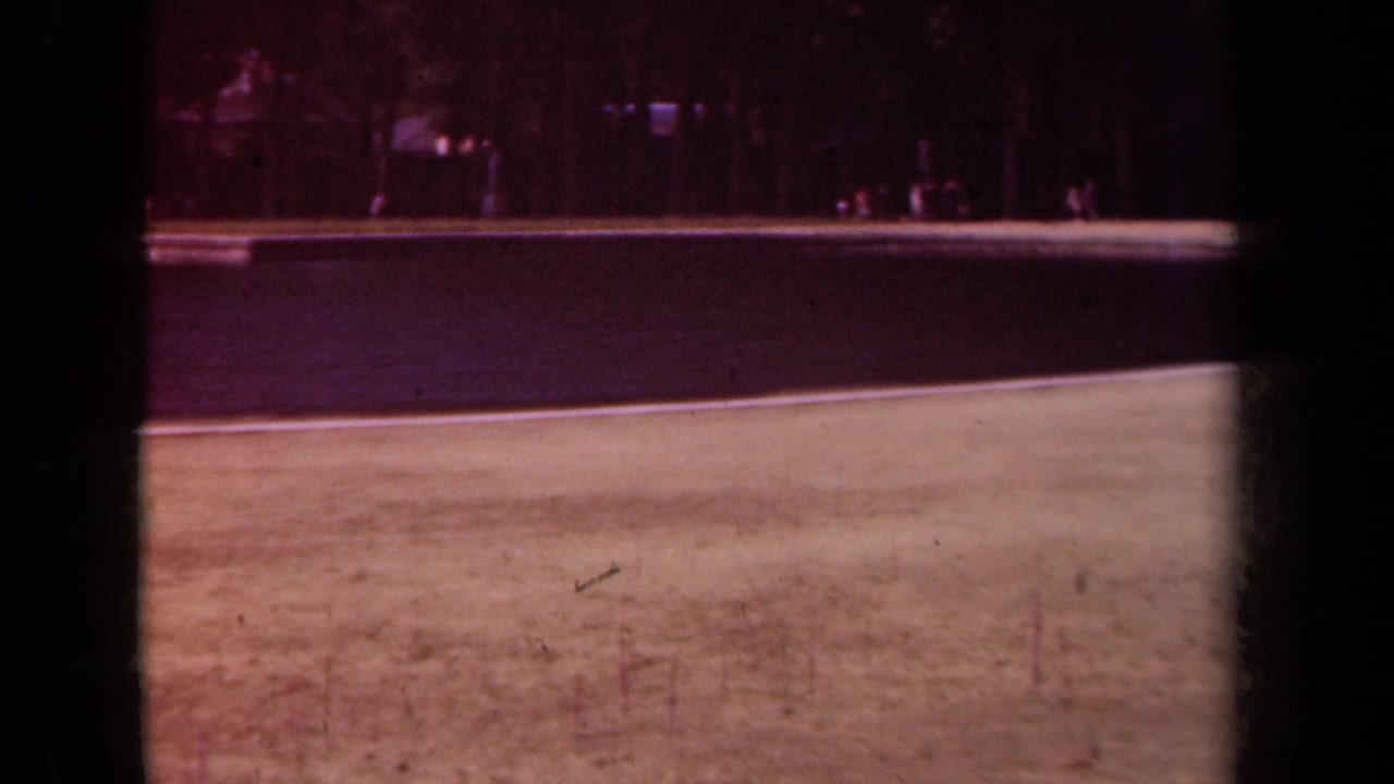 Park water and grass seen through dark film borders, with trees and small figures beyond.