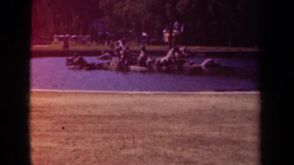 1939 Paris 8mm frame showing the fountain sculpture from a low, distant viewpoint, with water, stone edging, trees, and park visitors visible.