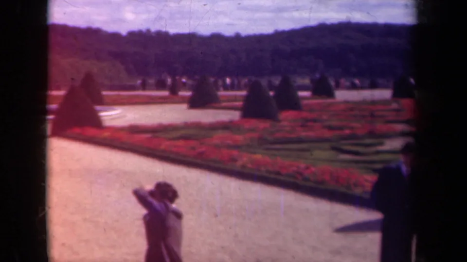 1939 Paris palace garden scene with clipped topiary, red flowerbeds, gravel paths, distant visitors, and a woman in the foreground.