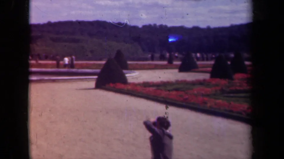 Handheld 1939 8mm view across a Paris palace garden path, red flowerbeds, clipped topiary, distant visitors, and a foreground woman.