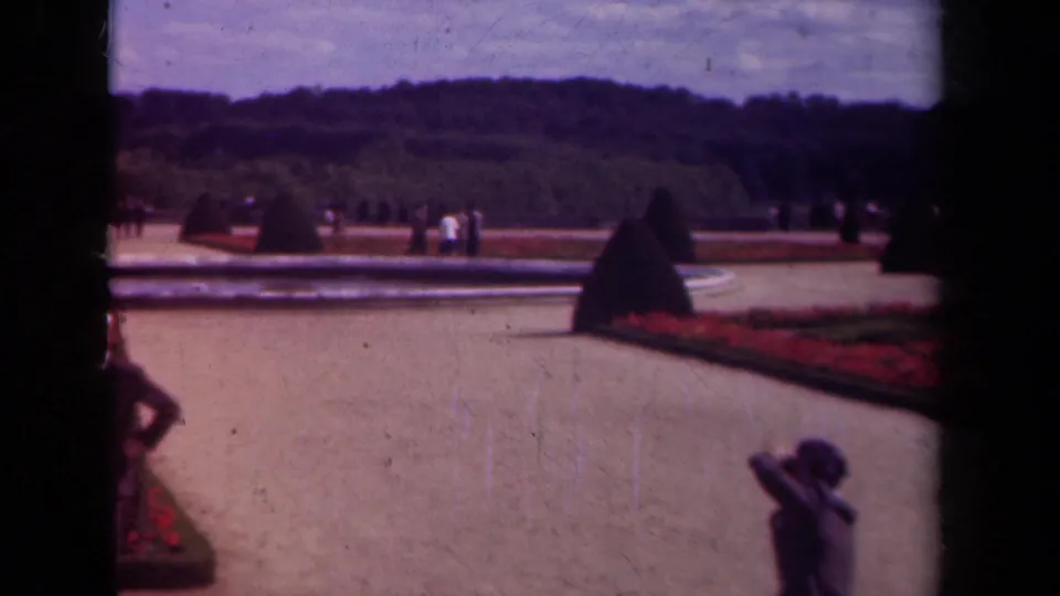 1939 Paris palace garden frame showing a broad path, red planting beds, clipped topiary, visitors in the distance, and a woman at lower right.