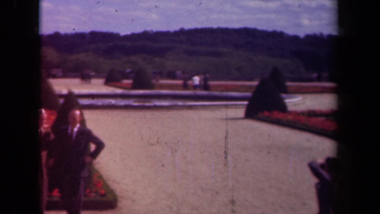 1939 Paris home-movie frame showing two men near red flowerbeds, clipped topiary, a pale reflecting pool, and distant garden visitors.