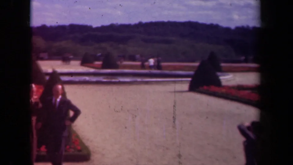 1939 Paris home-movie frame showing two men near red flowerbeds, clipped topiary, a pale reflecting pool, and distant garden visitors.