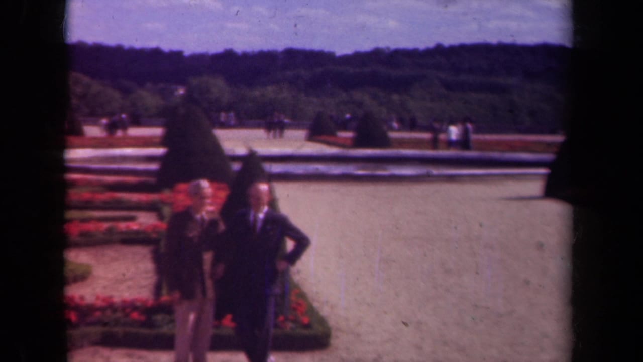 Two men in suits walk beside red flowerbeds and a reflecting pool in a formal garden.