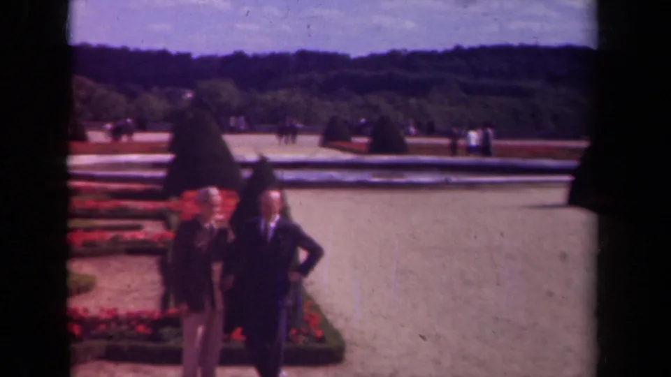 1939 Paris frame with two suited men crossing the foreground near geometric flowerbeds, topiary, and a long reflecting pool.