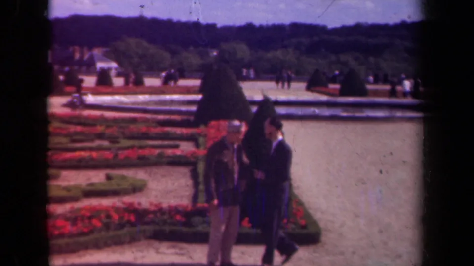 1939 Paris garden view showing two men near shaped topiary, red planting beds, a pale pool, and visitors scattered across the grounds.