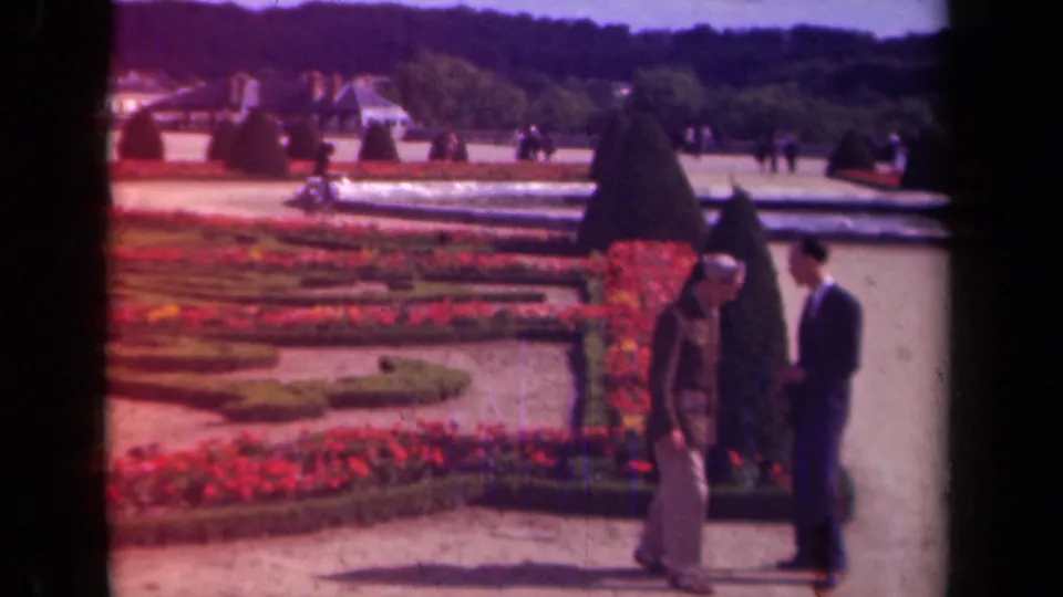 1939 Paris home-movie frame of two men paused by clipped topiary and red flowerbeds, with a reflecting pool and garden visitors in the distance.