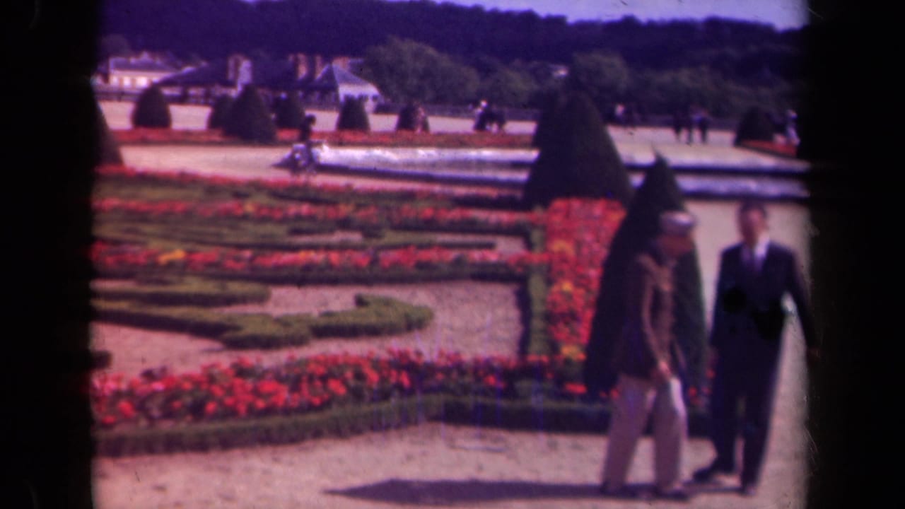 In metadata-supported 1939 Paris, two adults pass through a formal garden framed by dark 8mm film edges, red flowers, hedges, and topiary.