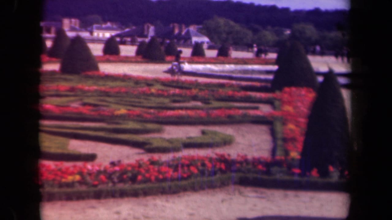 A 1939 Paris formal garden view shows red flower beds, geometric hedges, cone-shaped topiary, pale buildings, and scattered visitors in soft 8mm color.