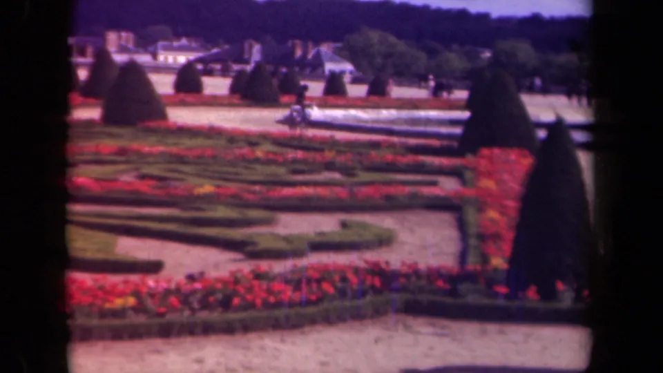 A 1939 Paris formal garden view shows red flower beds, geometric hedges, cone-shaped topiary, pale buildings, and scattered visitors in soft 8mm color.