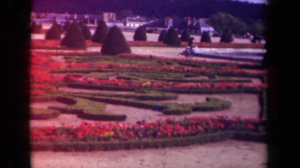 The garden is shown without close foreground figures, emphasizing red flowers, low clipped hedges, paths, topiary, and distant buildings in vintage 8mm color.