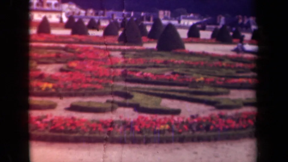 A late-1930s Paris garden view, supported by metadata, shows red flower beds, clipped hedges, topiary forms, distant visitors, and pale buildings through 8mm texture.