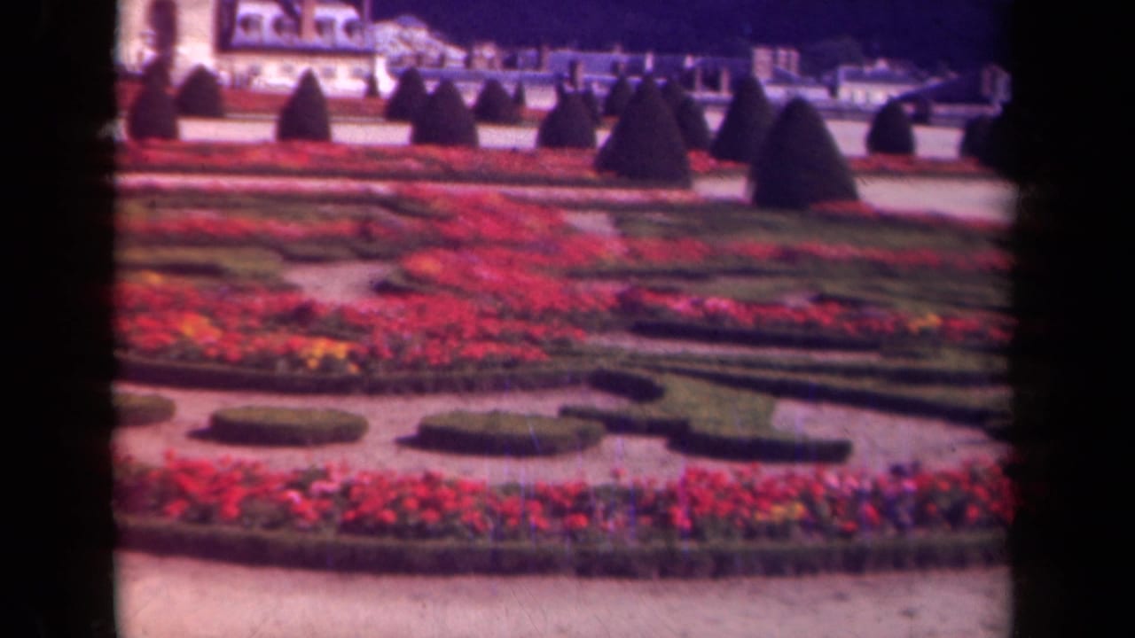 Formal garden with red flower beds, clipped hedges, topiary, and a classical building beyond.