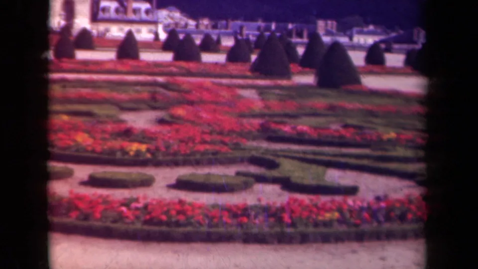 1939 Paris home-movie frame showing a formal garden with red flowers, shaped hedges, dark topiary, and a pale classical facade in the background.