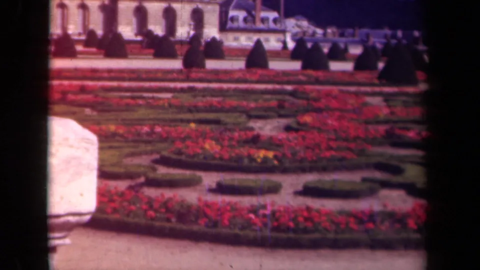 1939 Paris home-movie frame of a formal flower garden, with a pale foreground post, red beds, clipped hedges, topiary, and distant visitors.