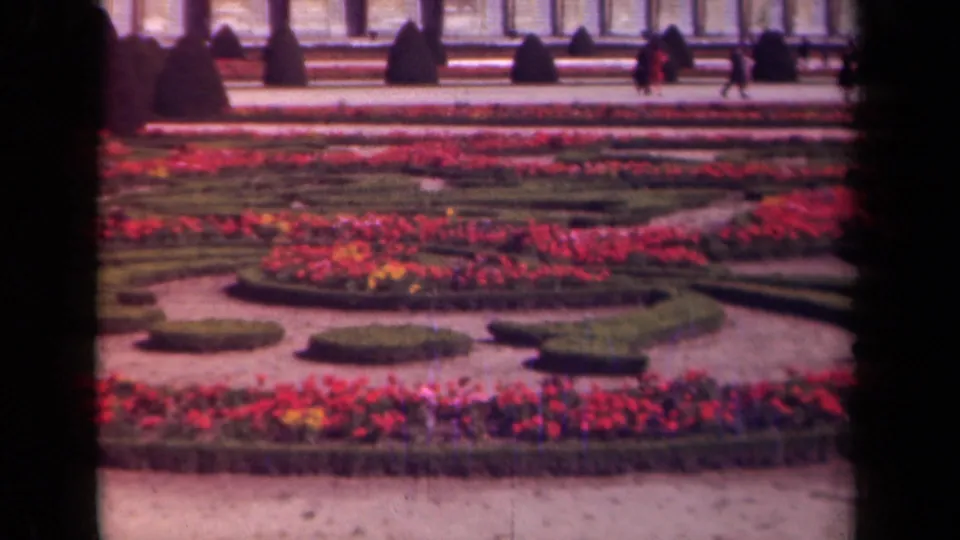 1939 Paris garden frame showing red flower beds, shaped hedges, gravel paths, dark topiary, and small distant visitors near a classical building.
