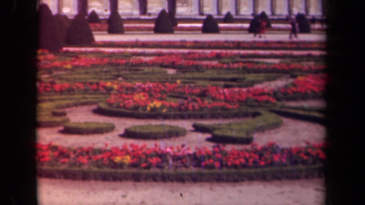 Formal flower beds and hedges before a large classical building with distant walkers.