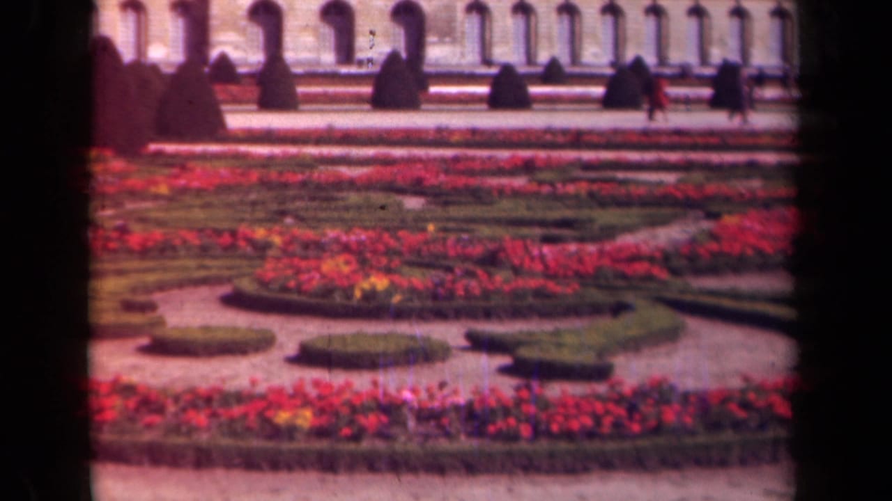 1939 Paris travel footage frame showing symmetrical flower beds and hedges below a long arched classical facade with distant pedestrians.
