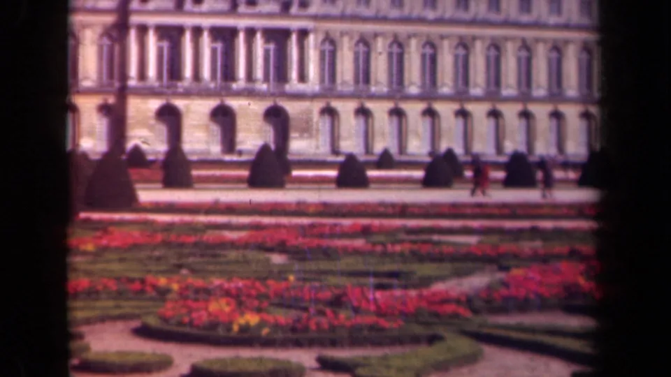1939 Paris home-movie frame with red flower beds, shaped hedges, and small visitor figures moving before a pale monumental building.
