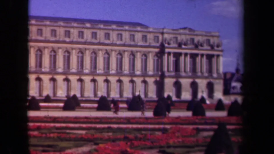 Soft 1939 8mm view of a Paris historic building and formal gardens, with small figures walking near clipped shrubs.