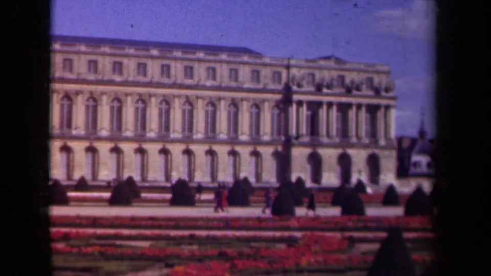 1939 Paris travel footage frame with red flowerbeds, clipped shrubs, a grand facade, and distant visitors in the garden.