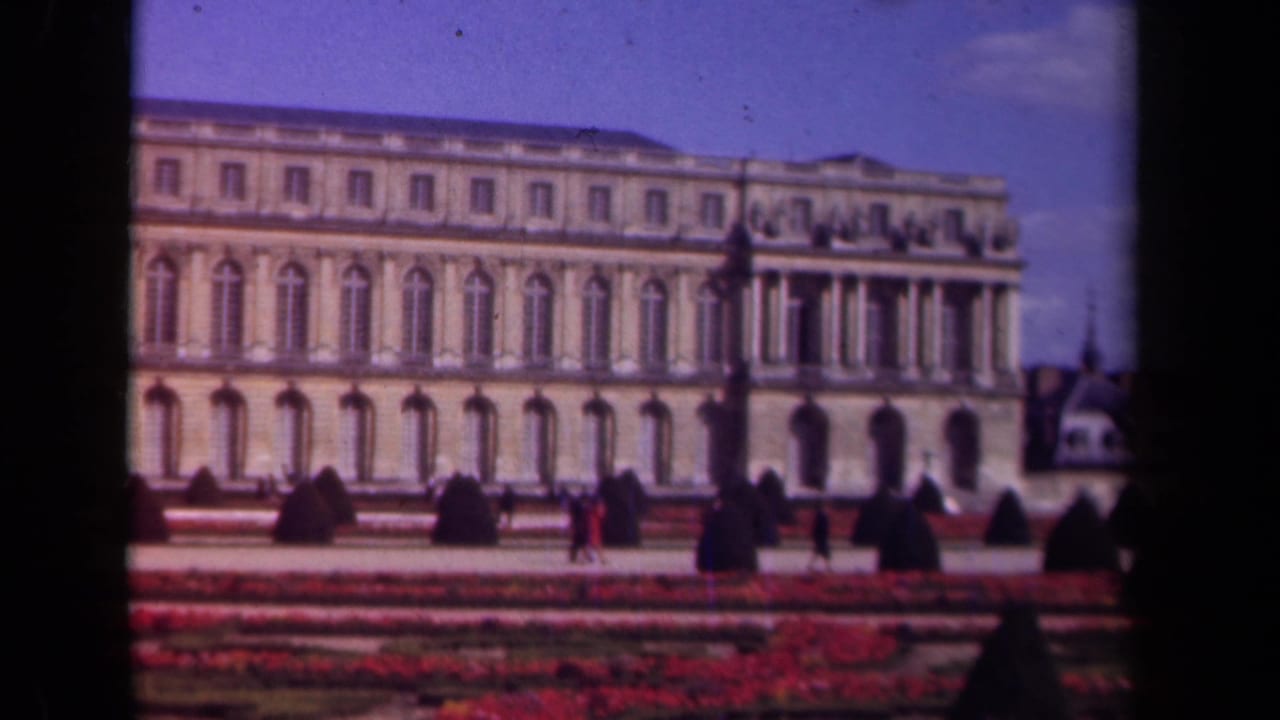 Classical stone building behind formal red flowerbeds with visitors crossing the grounds.
