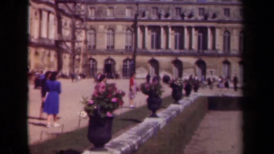 1939 Paris frame with a woman in a blue dress, flower planters, visitors, and a large historic stone building in the background.