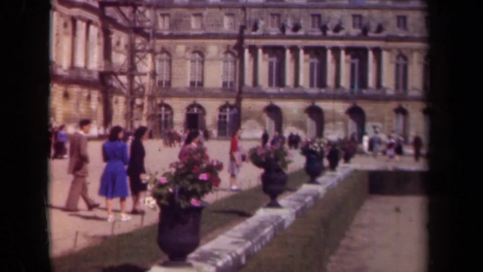 Paris, 1939 home-movie frame showing visitors in period dress crossing a palace courtyard with potted flowers and a formal stone facade.