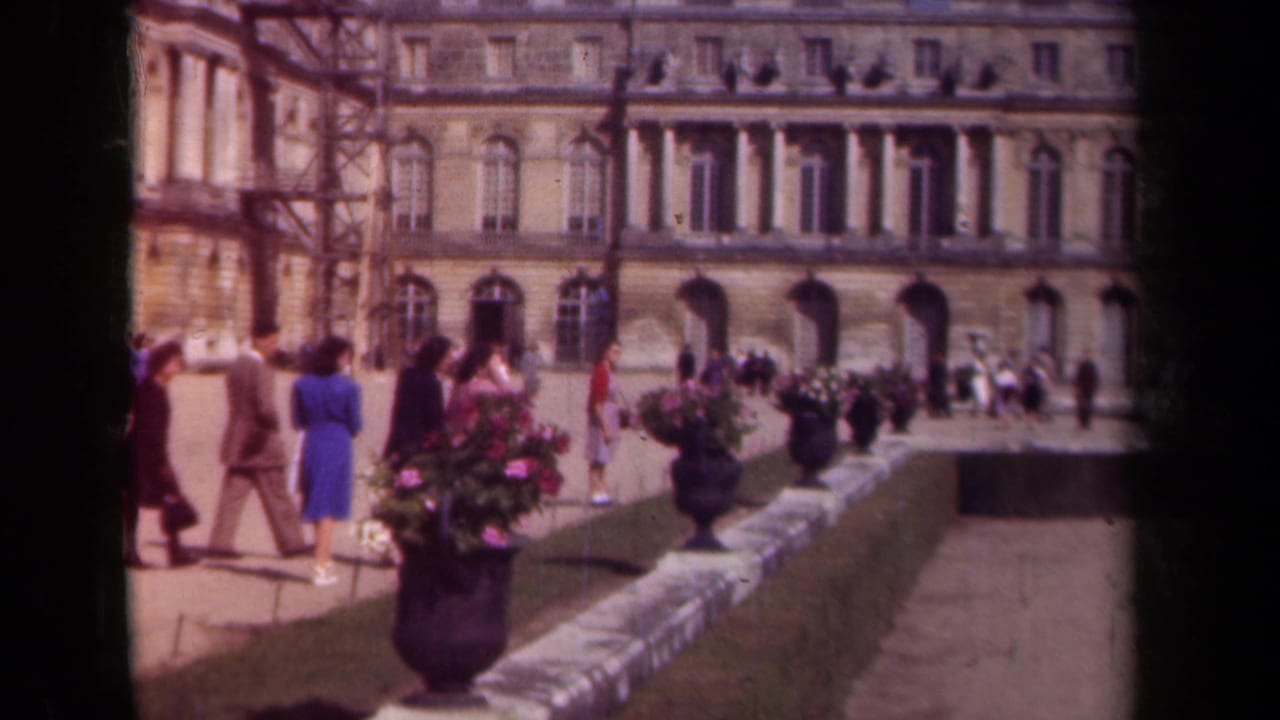 People stroll past flower planters in front of a formal stone building with arched openings.