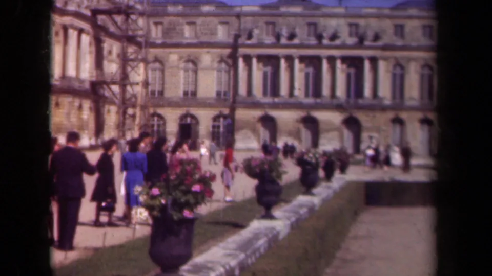 Paris, 1939 frame showing a small group near a flower planter and other visitors moving across a sunlit palace courtyard.
