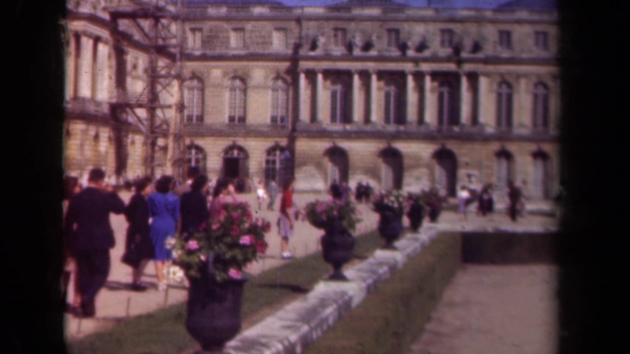 1939 Paris home-movie frame showing tourists in formal gardens near a large stone facade with flower urns and paths.