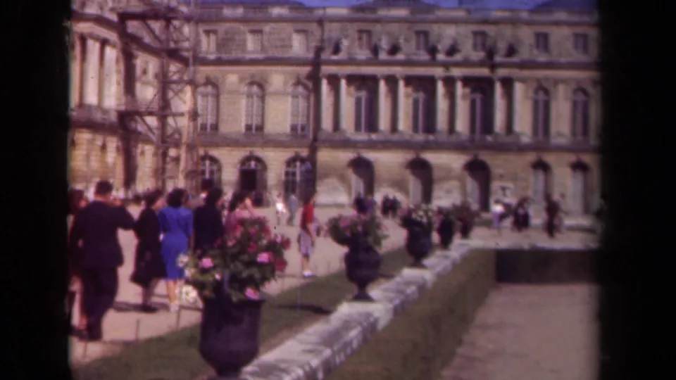 1939 Paris home-movie frame showing tourists in formal gardens near a large stone facade with flower urns and paths.