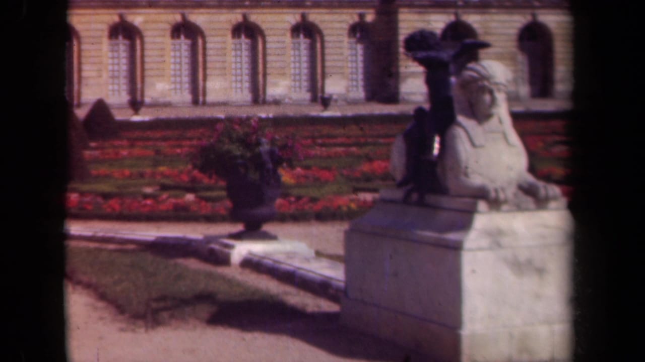 1939 Paris frame with a stone sphinx statue, red flowerbeds, urn planter, and arched windows in the background.