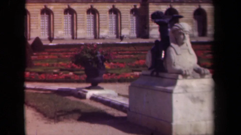 1939 Paris frame with a stone sphinx statue, red flowerbeds, urn planter, and arched windows in the background.
