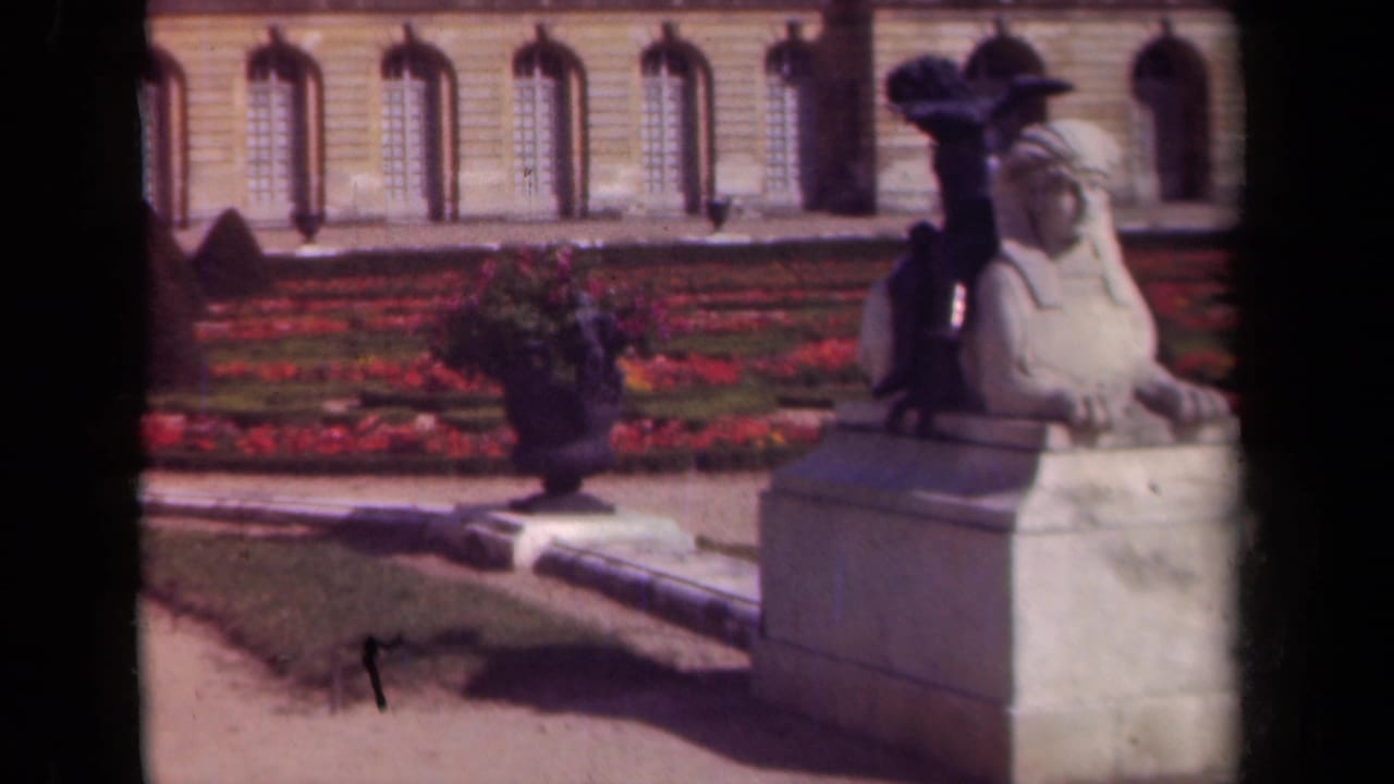 Stone sphinx statue beside a flower urn in a formal garden with a classical building behind it.