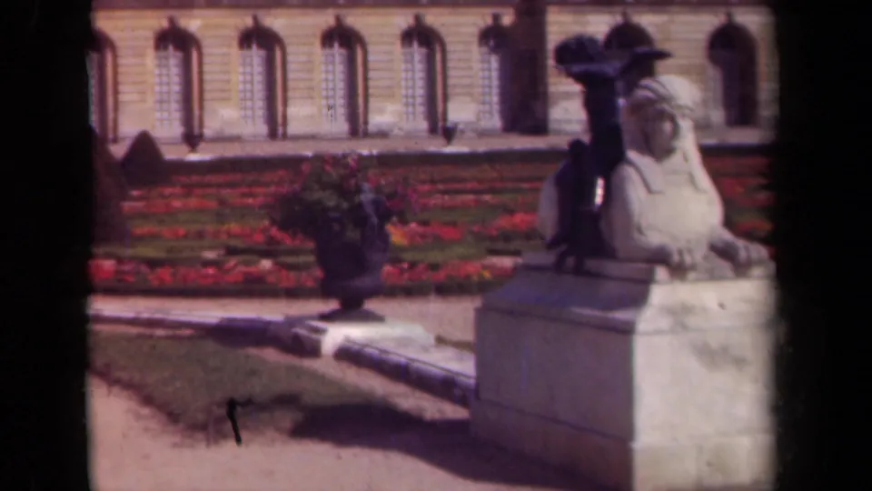 1939 Paris garden view with a stone sphinx statue, flower urn, red beds, lawn, and classical facade in soft 8mm color.