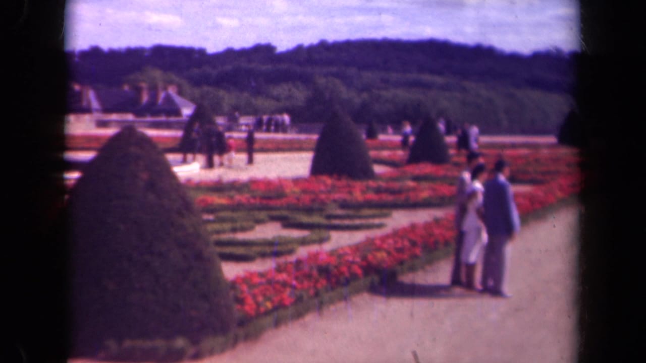 1939 Paris home-movie frame showing a couple near red flower beds and clipped cone topiary in a formal gravel garden.