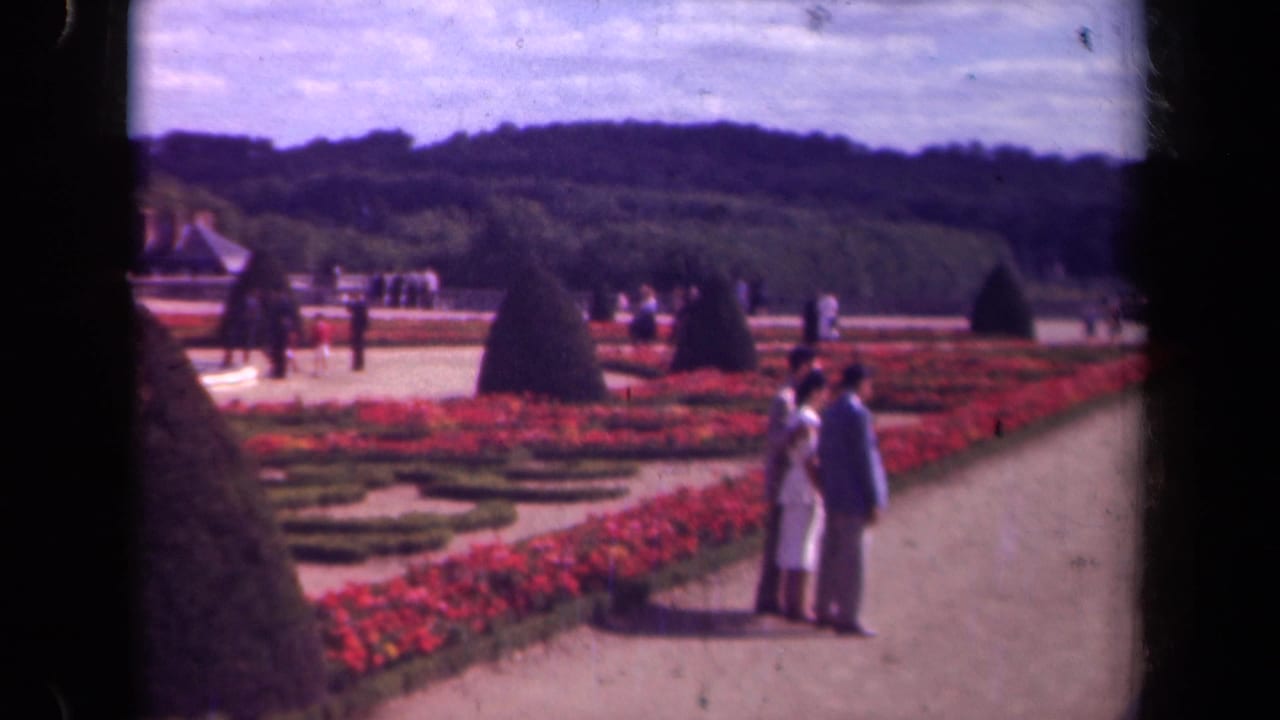 1939 Paris garden view with a couple in light clothing, red geometric planting, cone-shaped topiary, and distant visitors.