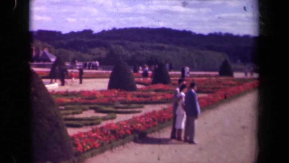1939 Paris garden view with a couple in light clothing, red geometric planting, cone-shaped topiary, and distant visitors.