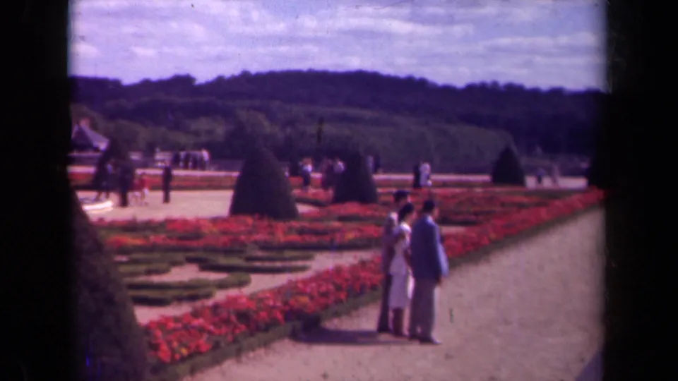 1939 Paris home-movie frame of visitors in a formal garden with red beds, clipped hedges, and cone-shaped topiary.