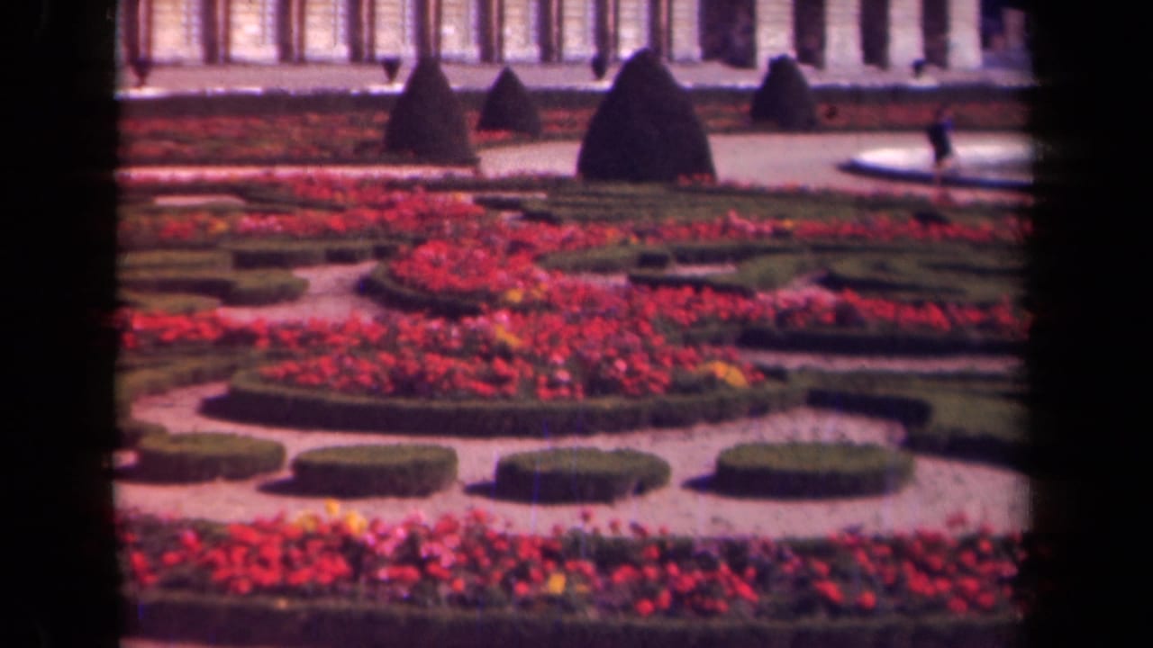 Formal garden with red flower beds, trimmed hedges, gravel paths, topiary, and a classical facade behind it.