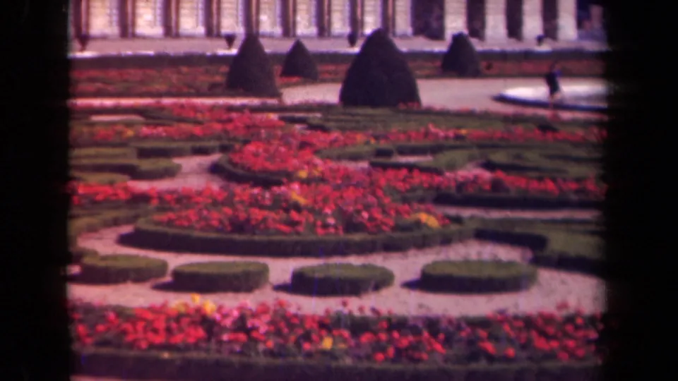 1939 Paris-area home-movie view of a formal garden with red flowers, clipped hedges, gravel paths, dark topiary, and a columned building facade.
