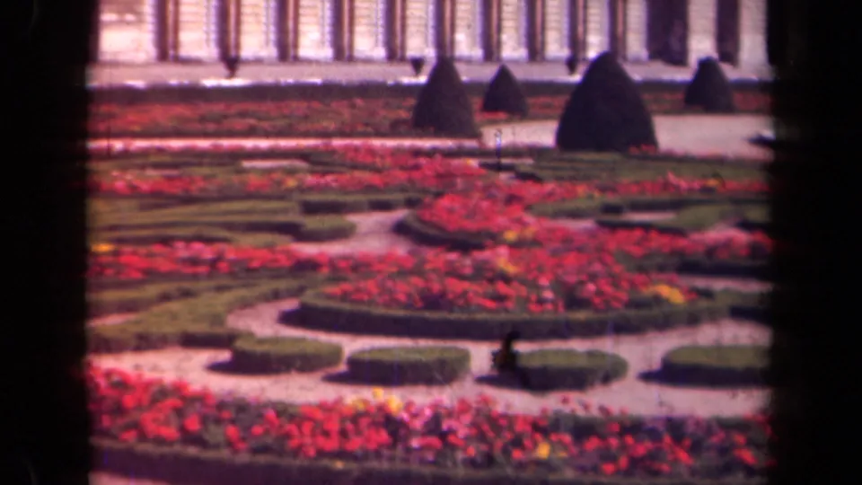 1939 home-movie frame of a formal Paris-area garden, with curving gravel paths, red flower beds, clipped hedges, topiary, and a grand facade.