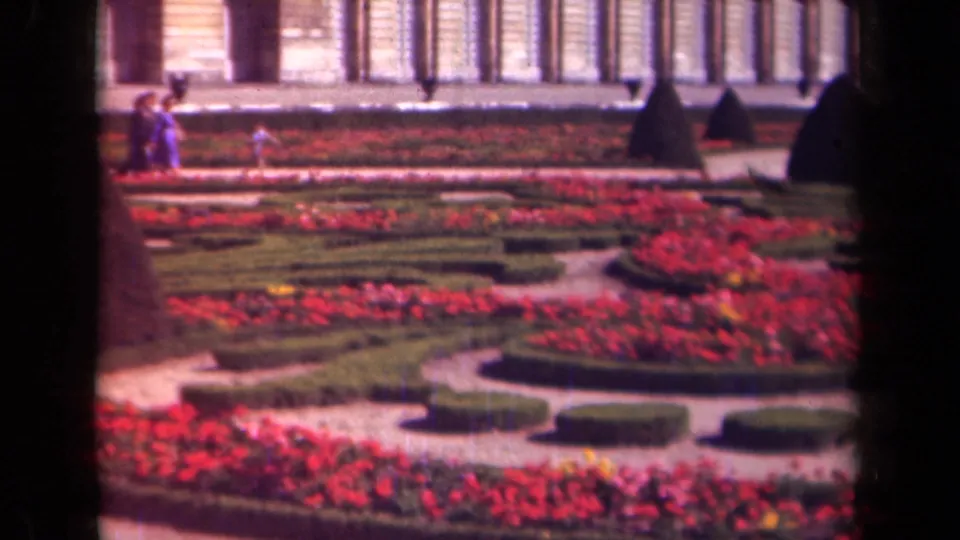 Paris-area 1939 garden view with distant visitors near red planting beds, low hedges, gravel paths, topiary, and a columned background facade.